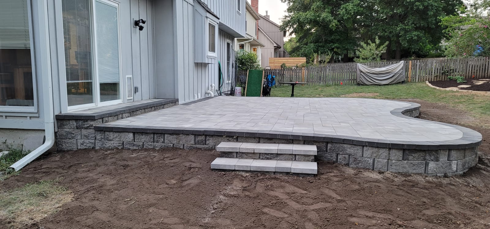 Raised gray paver patio with retaining wall blocks, curved border, and steps beside a Kansas City home.