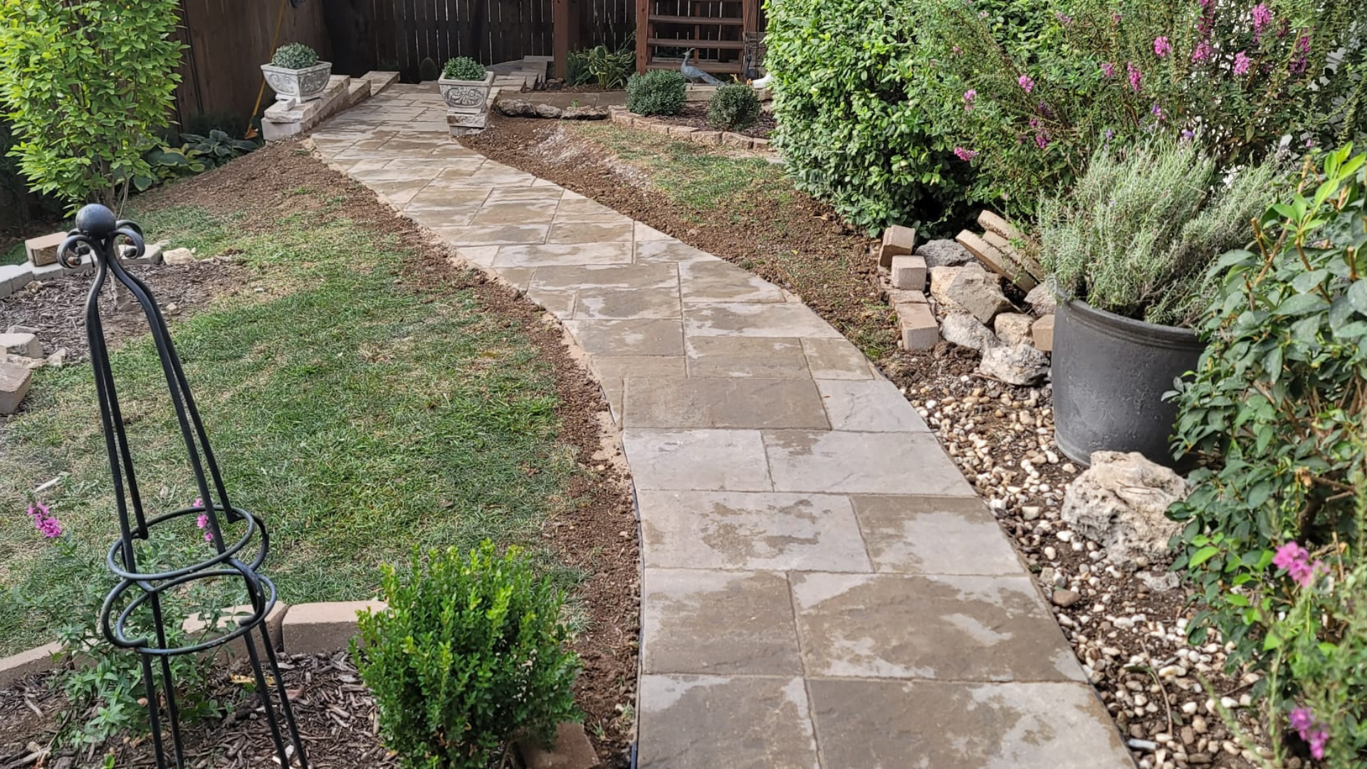 Backyard garden walkway with pavers, mulch beds, shrubs, and decorative rock.