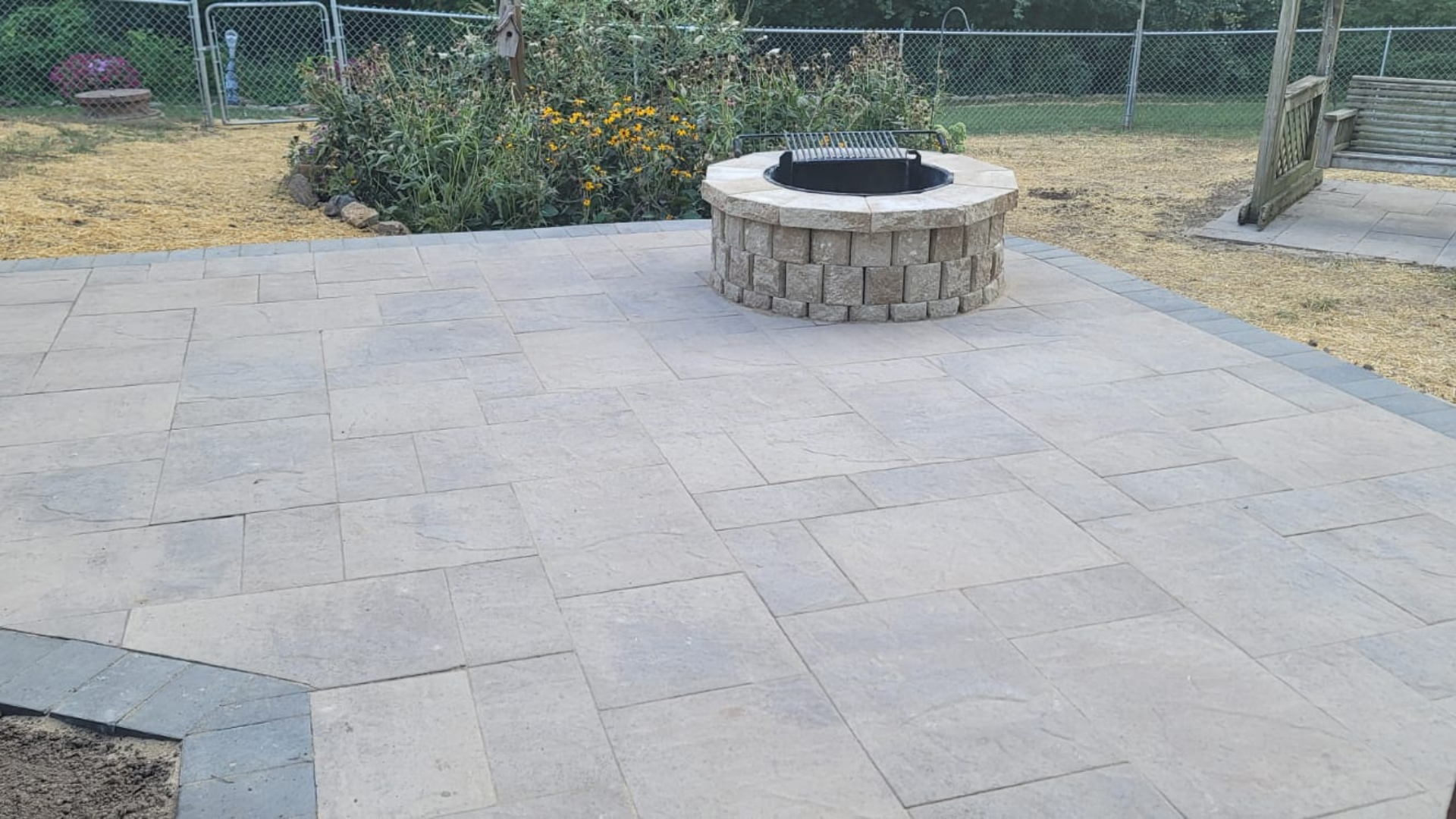 Large backyard paver patio with circular stone fire pit and garden bed.