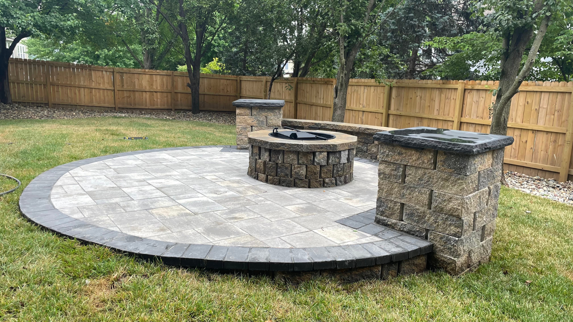 Backyard paver patio with circular fire pit, stone pillars, and seating wall.