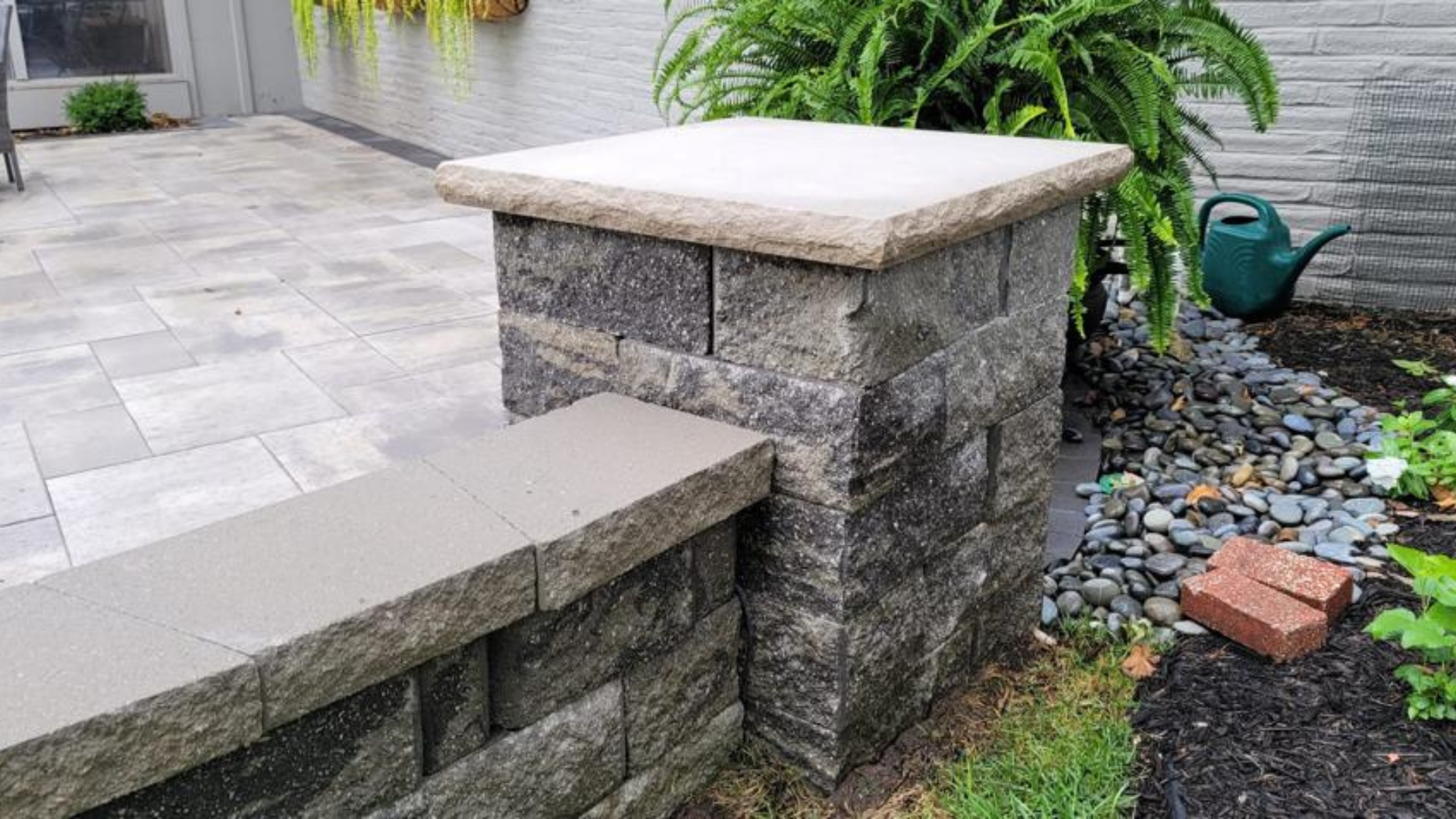 Close-up of stone patio pillar and seating wall beside pavers and landscape bed.