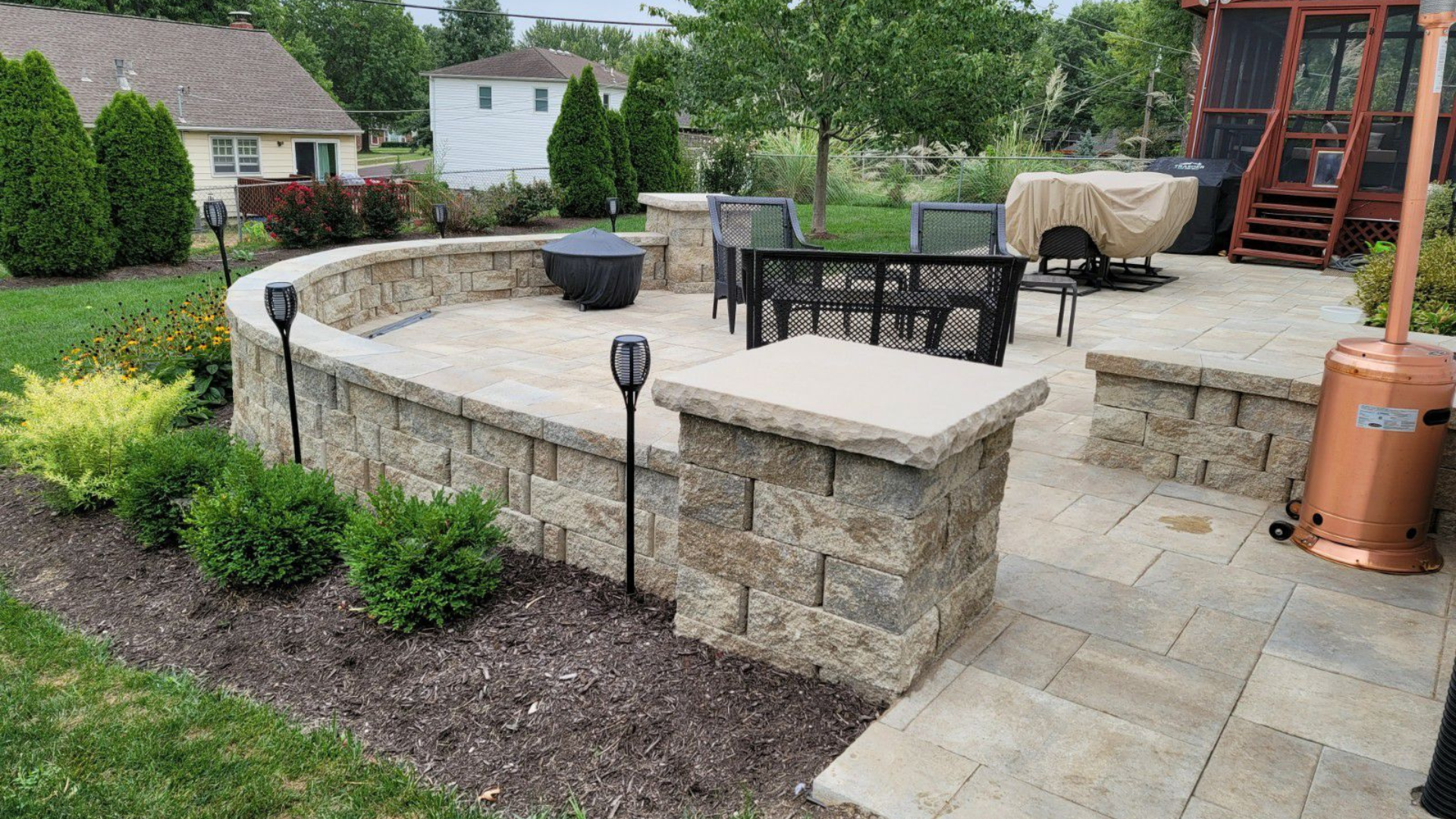 Raised paver patio with stone retaining wall, outdoor seating, and landscape beds.
