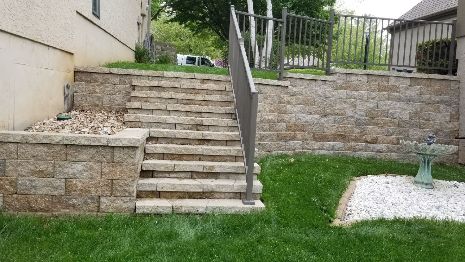 Retaining wall with stone stairway, metal railing, lawn, and rock bed.