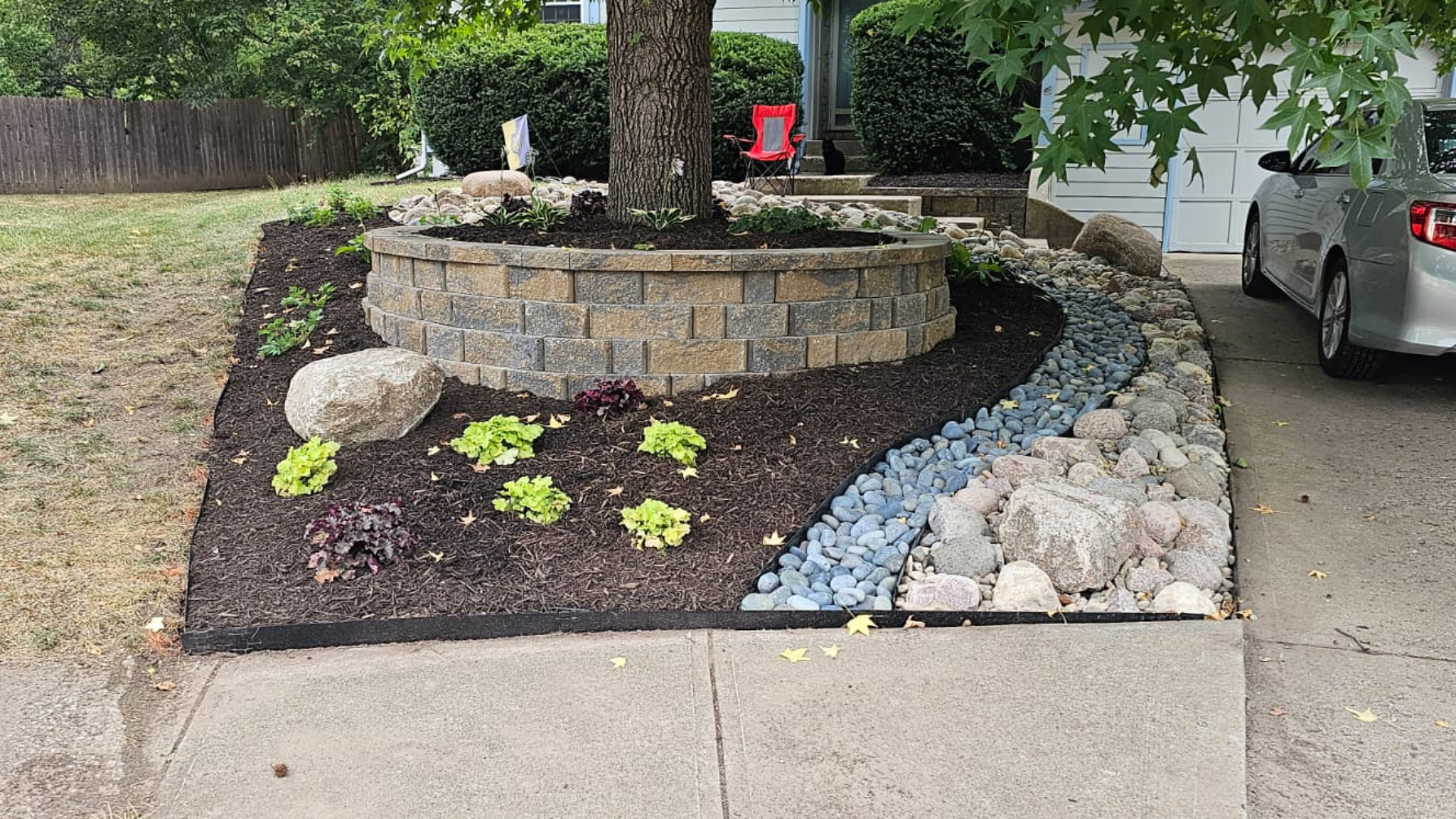 Tree ring retaining wall with mulch, plants, boulders, and decorative dry creek bed.