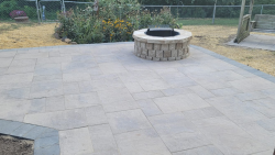 A spacious paver patio with fire pit creates room for outdoor entertaining. Large backyard paver patio with circular stone fire pit and garden bed.