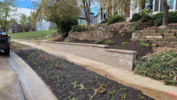 A large retaining wall improves stability and appearance on a sloped property. Large retaining wall with sidewalk, mulch bed, and hillside landscaping.